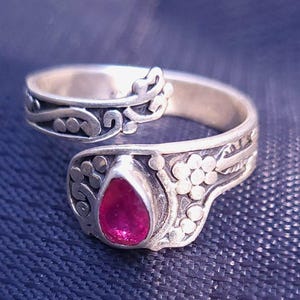 Peut inclure: Une bague argentée avec un design ouvert, ornée d'une pierre précieuse rouge vif en forme de poire. La bague est ornée de motifs floraux et de volutes complexes. La bague est posée sur un fond texturé bleu foncé.