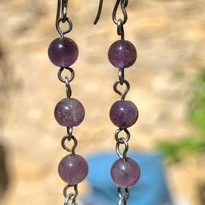 Puede incluir: Pendientes colgantes con una serie de cuentas de amatista moradas unidas por eslabones de metal plateado. Los pendientes tienen un cierre de gancho y terminan con una cuenta más grande y multicolor. Aproximadamente 6 cm de largo.