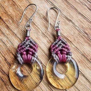 Peut inclure: Paire de boucles d'oreilles faites à la main. Chaque boucle présente un pendentif circulaire transparent de couleur ambre avec un effet de grain de bois. La partie supérieure est en macramé gris et magenta, avec des crochets d'oreilles argentés.