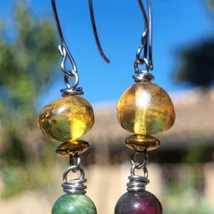 Pode incluir: Um par de brincos pendurados com ganchos prateados. Cada brinco apresenta uma grande conta translúcida cor de âmbar, um pequeno disco dourado e um design de fio enrolado. Uma conta é verde, a outra vermelho escuro.
