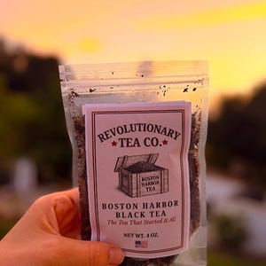 May include: A clear plastic bag of Boston Harbor Black Tea held in front of a sunset. The label reads "Revolutionary Tea Co." with an illustration of a wooden chest. The text "Boston Harbor Black Tea, The Tea That Started It All" is also visible. Net weight is 4 oz.