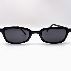 May include: Black sunglasses with a rectangular frame and dark lenses. The sunglasses have a sleek, modern design. The arms are thin and extend to the temples. The frames are made of a glossy material.