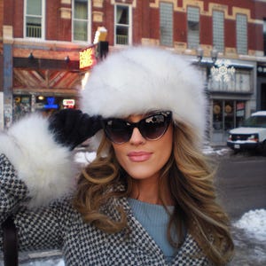 Könnte beinhalten: Eine Frau trägt einen weißen Pelzhut, eine schwarze Sonnenbrille und einen schwarz-weiß gemusterten Mantel. Der Hut und die Handschuhe haben einen weißen Pelzbesatz. Im Hintergrund ist eine Stadtstraße mit Backsteingebäuden und Schnee zu sehen.