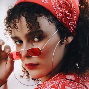 May include: A woman wearing a red bandana with a paisley pattern, red-tinted rectangular sunglasses, and large gold hoop earrings. Her lips are painted red, and she has red eyeshadow. She is also wearing a red paisley shirt.