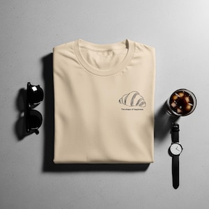 Puede incluir: Una camiseta beige con un dibujo lineal negro de un cruasán y el texto "The shape of happiness". Gafas de sol negras, un reloj de pulsera y un vaso de café helado también están en la imagen.