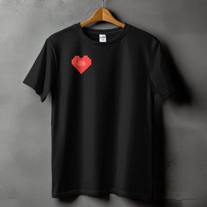 以下が含まれることがあります： 胸に赤いピクセル化されたハートのグラフィックが付いた黒いTシャツ。ハートには白い文字が含まれています。シャツは灰色の壁を背景に木製のハンガーに吊るされています。