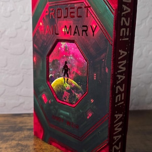 May include: A hardcover book titled Project Hail Mary by Andy Weir with a red and green cover featuring a central octagonal window showing a silhouette on a planet. The spine displays the repeated text AMAZE! AMAZE! AMAZE! in white lettering.
