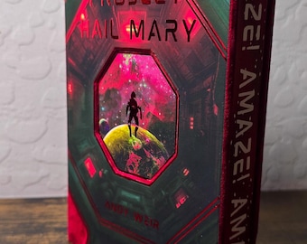 May include: A hardcover book titled Project Hail Mary by Andy Weir with a red and green cover featuring a central octagonal window showing a silhouette on a planet. The spine displays the repeated text AMAZE! AMAZE! AMAZE! in white lettering.