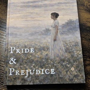 Op de afbeelding: Een boek met de titel "Pride & Prejudice". De omslag toont een vrouw in een witte jurk in een veld. De titel is wit. De achtergrond toont een bewolkte lucht en gras.