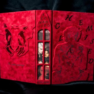 Op de afbeelding: Een rood boek met de titel "A Hemised Claded" in reliëf op de kaft. De kaft heeft uitgesneden ontwerpen van handen en een silhouet van een persoon. Een raam onthult twee mensen binnenin.