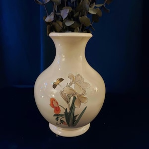 May include: Cream-colored ceramic vase with a wide base and a flared top. The vase features a floral design with white irises, orange flowers, and a butterfly. The vase is filled with dried green leaves.