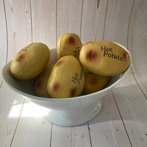 May include: A white, pedestal serving bowl filled with several yellow, potato-shaped toys. Each toy has a brown spot and the words "Hot Potato" printed on it. The bowl is set against a light wood-grain background.