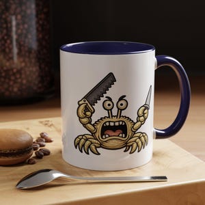 May include: A white ceramic mug with a dark blue interior and handle. The exterior features a graphic illustration of a tan crab with an aggressive expression, holding a hand saw in one claw and a small knife in the other claw, centered on the mug.