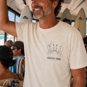 May include: A cream-colored short-sleeve t-shirt featuring a black line-art illustration of a crab holding a knife and fork on the left chest area, with the text "BEACH TIME" printed in black capital letters directly below the crab graphic.