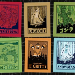 May include: Six colorful art prints featuring cryptids. The prints include the Jersey Devil, Bigfoot, Godzilla, Old Greeny, It Gritty, and the Abominable Snowman. Each print has a unique color scheme and stylized illustration.