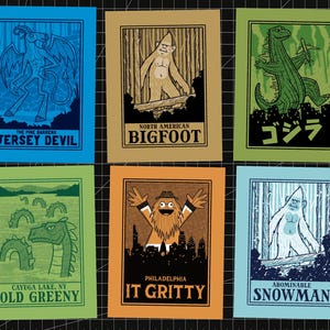 May include: Six rectangular prints featuring stylized illustrations of mythical creatures. The designs include the Jersey Devil, Bigfoot, Godzilla with Japanese text, Old Greeny, It Gritty in Philadelphia, and the Abominable Snowman, each with bold borders.