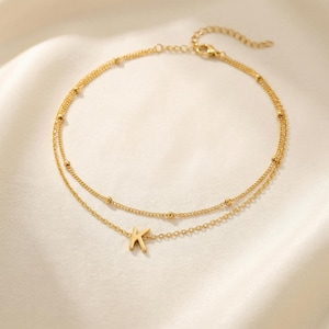 May include: A gold-colored, double-layered anklet with a small letter "K" charm. The anklet features a delicate chain design with small, evenly spaced beads. The jewelry is displayed on a white, silky surface.