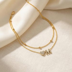 May include: A gold-colored, double-layered anklet with a delicate chain design. The anklet features small, round beads and a heart-shaped charm with the letter "M" in a sparkling, crystal-like material. The anklet is displayed on a white, satin-like fabric.
