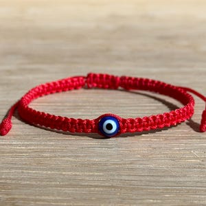 May include: Red woven bracelet featuring a blue and white evil eye bead. The bracelet is adjustable with sliding knots. The evil eye bead is a protective amulet, often worn for good luck and warding off negative energy.