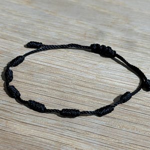 May include: A black knotted bracelet with a sliding knot closure. The bracelet features several evenly spaced knots along its circumference. The bracelet is made of a dark, possibly nylon, cord. The background is a light brown wooden surface.