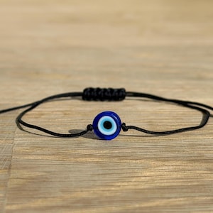 May include: A black string bracelet featuring a blue and white evil eye charm. The charm is round with a dark blue outer ring, a light blue inner ring, and a black center. The bracelet has adjustable sliding knots.