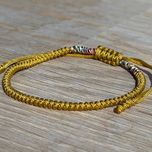 以下が含まれることがあります： マルチカラーのアクセントが付いた、金色の織りブレスレット。 ブレスレットは、中央と端に結び目のあるデザインで、しっかりと織られたコードで作られています。 背景は、明るい茶色の木製の表面です。