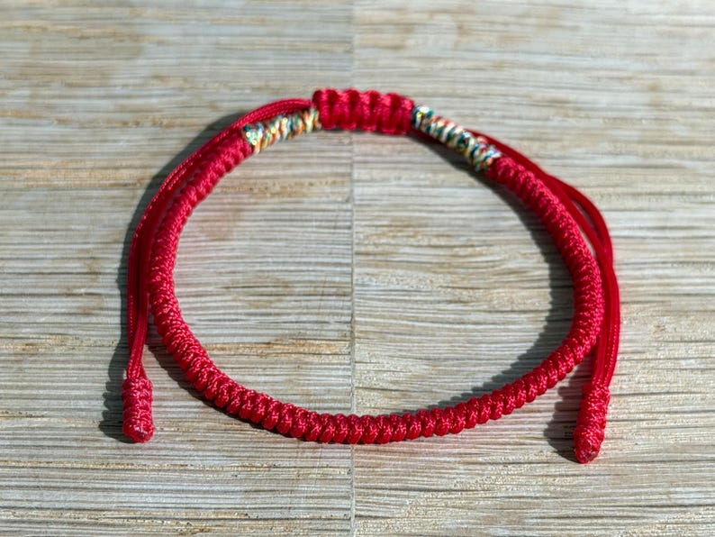 以下が含まれることがあります： 赤い編み込みブレスレット。ブレスレットは、カラフルなアクセントが付いた装飾部分があります。ブレスレットは、明るい茶色の木製の表面に表示されています。