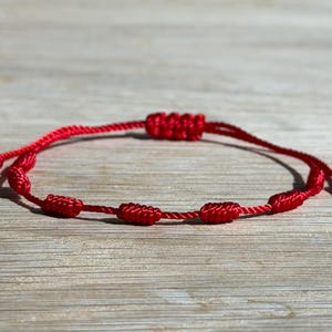 May include: A red string bracelet with multiple knots and an adjustable sliding knot closure. The bracelet is made of a thin, woven cord and is displayed on a light-colored wooden surface. The bracelet is a simple, minimalist design.