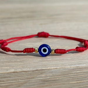 May include: Red string bracelet featuring a blue and white evil eye charm. The bracelet has a sliding knot closure for adjustable sizing. The charm is a deep blue with a white and black center, and gold-colored beads.