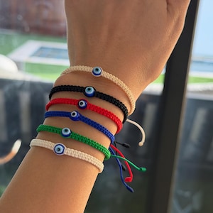 May include: A collection of six woven bracelets in various colors, including cream, black, red, blue, and green. Each bracelet features a small, round, blue and white evil eye charm. The bracelets are displayed on a wrist.