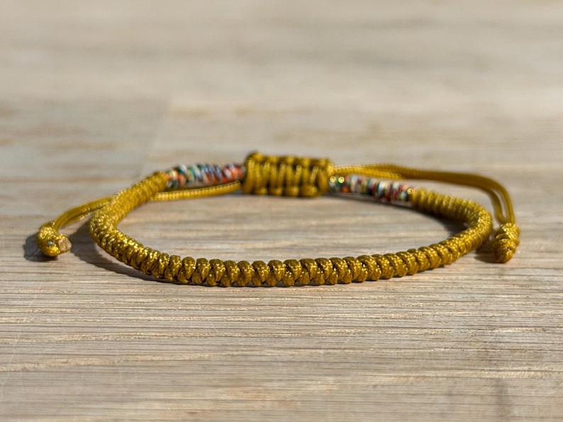 以下が含まれることがあります： ゴールドカラーの編み込みブレスレット。スライドノット留め。 ブレスレットは、詳細な編み込みデザインとノットの近くにカラフルなアクセントが特徴です。 ブレスレットは調節可能で、木製の表面に置かれています。