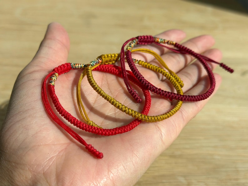 以下が含まれることがあります： 赤と金の3つの手作りのブレスレットが手のひらに展示されています。 ブレスレットは織り込まれたコードで作られ、装飾的な金属のアクセントが特徴です。 鮮やかなアクセサリーです。