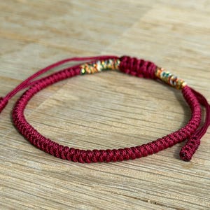 May include: A burgundy braided bracelet with adjustable cords. The bracelet features a decorative section with gold, green, and orange accents. The bracelet is displayed on a wooden surface.