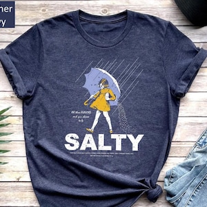 A navy blue t-shirt with a graphic design depicting a person holding an umbrella and sprinkling salt on the ground, accompanied by the text "SALTY" and a Bible verse reference.
