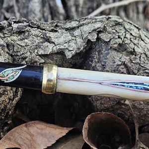 May include: A handcrafted pen with a dark wood barrel, accented with copper wire and a gold-colored band. The pen features a decorative tip with blue and turquoise accents, and a cream-colored body with a blue and white design.