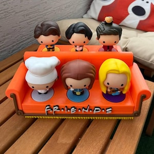 Könnte beinhalten: Ein orangefarbenes Miniatursofa beherbergt sechs Cartoon-Figuren, die jeweils einen Charakter aus der Fernsehserie "Friends" darstellen. Die Figuren haben unterschiedliche Frisuren und Outfits. Auf dem Sofa steht das Wort "FRIENDS".