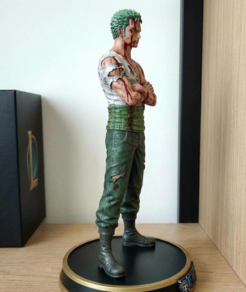 Op de afbeelding: Een beeldje van een mannelijk personage met groen haar en meerdere wonden. Hij draagt een gescheurd wit shirt, een groene broek en zwarte laarzen. Het beeld staat op een zwarte basis met gouden rand.