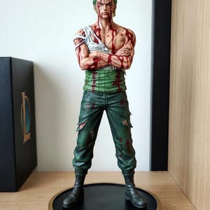 Op de afbeelding: Gedetailleerd beeldje van een personage met groen haar, gekleed in een gescheurd wit shirt, groene broek en zwarte laarzen. De figuur heeft meerdere rode, bloedachtige markeringen. De figuur staat op een zwart-gouden basis.