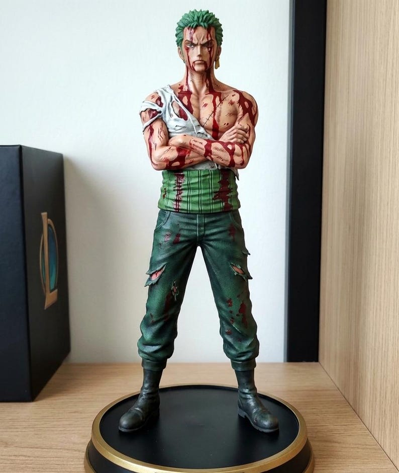 Op de afbeelding: Gedetailleerd beeldje van een personage met groen haar, gekleed in een gescheurd wit shirt, groene broek en zwarte laarzen. De figuur heeft meerdere rode, bloedachtige markeringen. De figuur staat op een zwart-gouden basis.