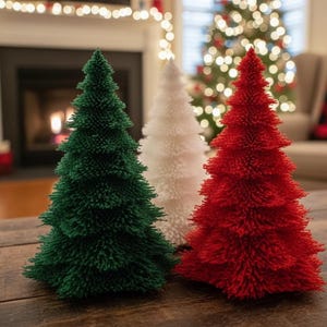 May include: Three decorative Christmas trees in green, white, and red. The trees are made of a textured material and are arranged on a wooden surface. The background includes a fireplace and a Christmas tree.