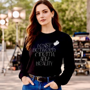 May include: Black long-sleeve shirt with the text "I Exist Between Cinema and Reality" in white. A white camera graphic is above the text. The shirt is paired with blue jeans and a black belt.