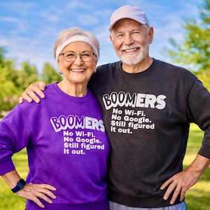 May include: A smiling senior couple wearing matching sweatshirts with the word "BOOMERS" in white block letters. The sweatshirts also read "No Wi-Fi. No Google. Still figured it out." The woman wears a purple sweatshirt, and the man wears a dark gray one.