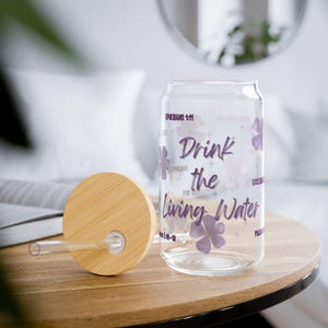 May include: A clear glass can-shaped drinking cup with a bamboo lid and straw. The cup features the text "Drink the Living Water" in purple script, along with floral and star designs. The cup is on a wooden table.