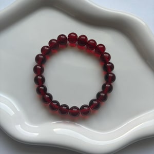 May include: A bracelet made of dark red, translucent beads. The bracelet is circular and sits on a white, wavy-edged plate. The beads are evenly sized and strung together, creating a simple and elegant accessory.