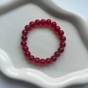 May include: A red beaded bracelet made of round, translucent beads. The bracelet is presented on a white, scalloped-edge plate. The beads are a deep, rich red colour.