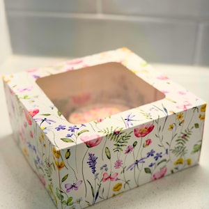 May include: A square bakery box with a clear window, decorated with a watercolor floral pattern. The design features pink, purple, yellow, and blue flowers, butterflies, and dragonflies on a white background. The box is designed to hold baked goods.