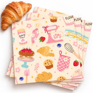 May include: A stack of square paper napkins featuring a repeating pattern of baking supplies including a pink stand mixer, whisk, oven mitt, and various pastries. A golden-brown croissant rests on the top left corner of the napkins.