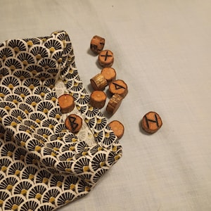 May include: A set of wooden rune stones with etched symbols, scattered beside a patterned fabric bag. The bag features a repeating fan-like design in black, brown, and gold. The runes are made of natural wood.