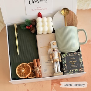 May include: A gift box with a green theme, including a green mug, a candle, a notebook, a pen, a nutcracker, tea, chocolate, and dried orange slices. The box has a card that reads "Christmas" and "From, The Harrington Family."