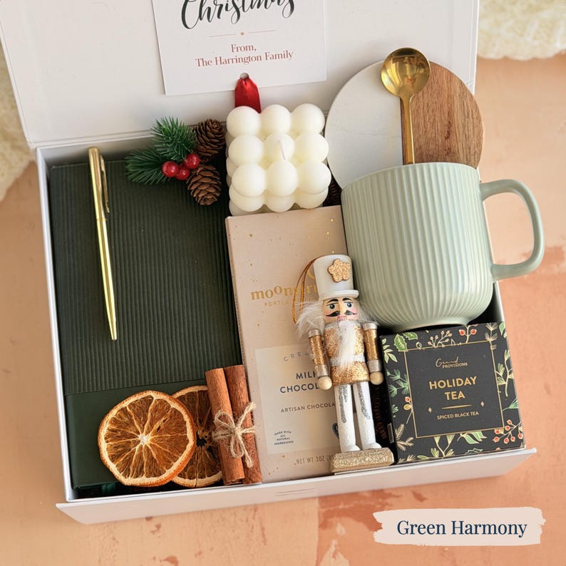 May include: A gift box with a green theme, including a green mug, a candle, a notebook, a pen, a nutcracker, tea, chocolate, and dried orange slices. The box has a card that reads "Christmas" and "From, The Harrington Family."